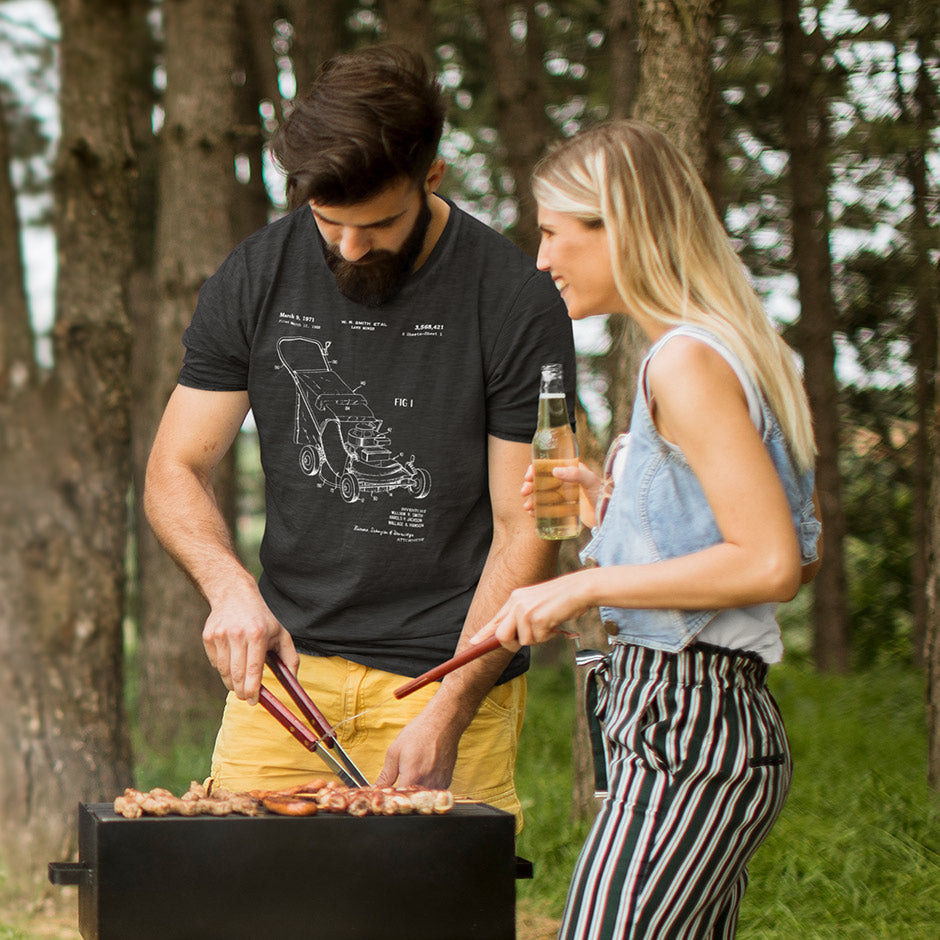 man wearing a BBQ shirt while standing by the grill. The mens tee features a 1971 patent drawing of a push lawn mower.