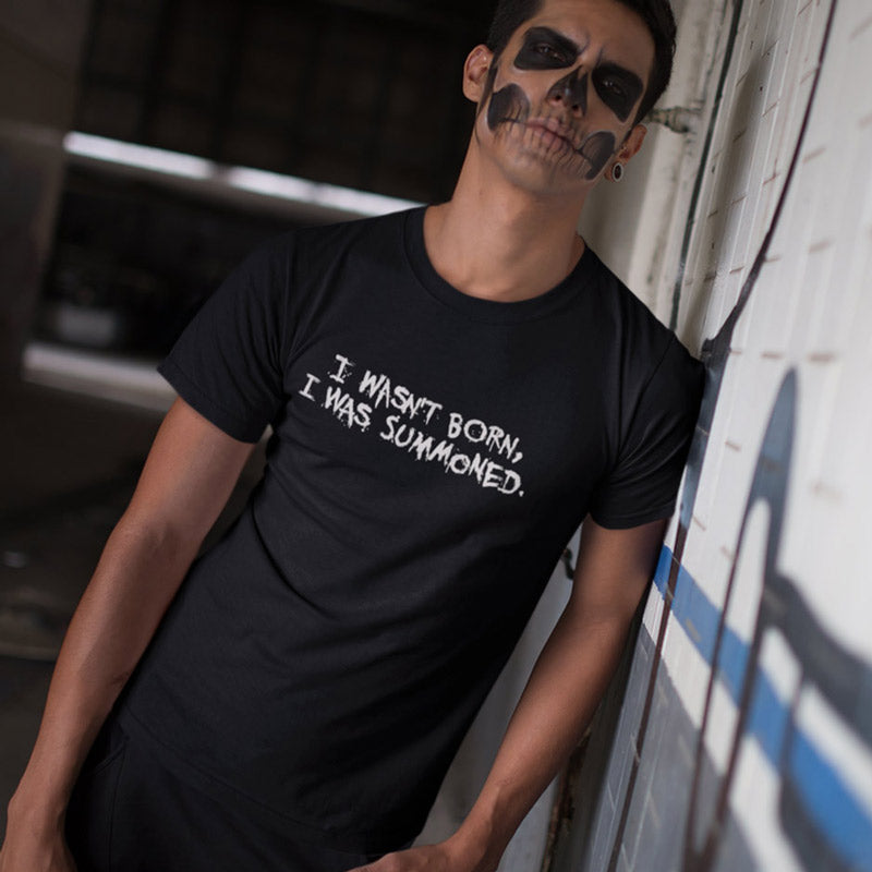 Guy leaning against a wall wearing a black halloween shirt that reads I wasn't born, I was summoned.