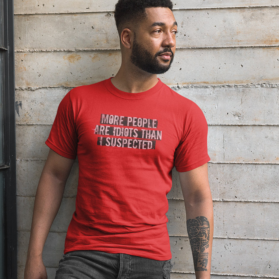 Man standing by wall wearing a red offensive t shirt that reads More people are idiots than I suspected. Angry Shirts design by Dodo Tees.