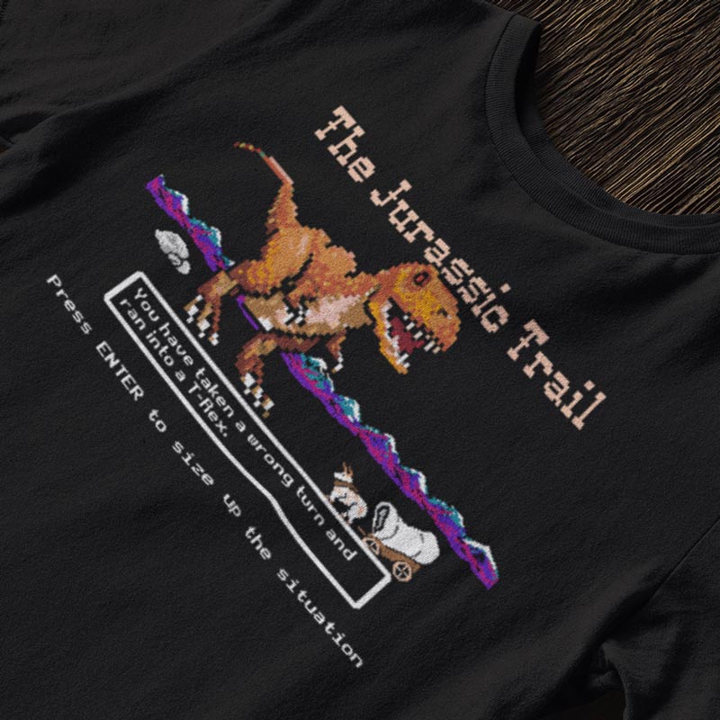 close up of the Jurassic Trail nerd shirt featuring The funny shirt features an 8 bit t rex dinosaur attacking an ox and wagon.