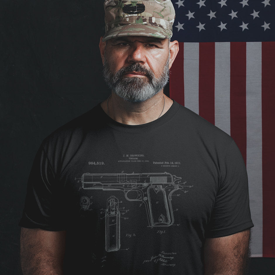 Military man in front of an American flag wearing a patriotic shirt featuring the 1911 pistol patent. Our side-seamed our firearms shirts are as enduring as the gun it celebrates.