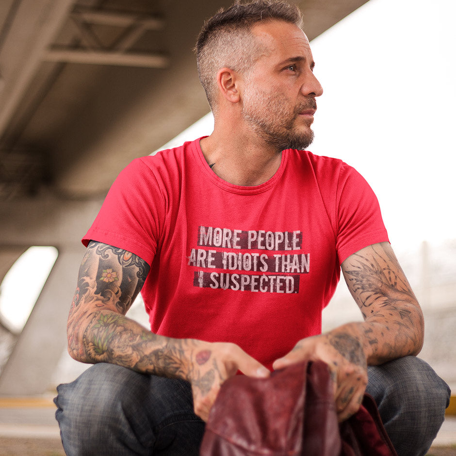 Man sitting on curb wearing a red sarcastic tee shirt that reads More people are idiots than I suspected. Funny Shirt design by Dodo Tees.