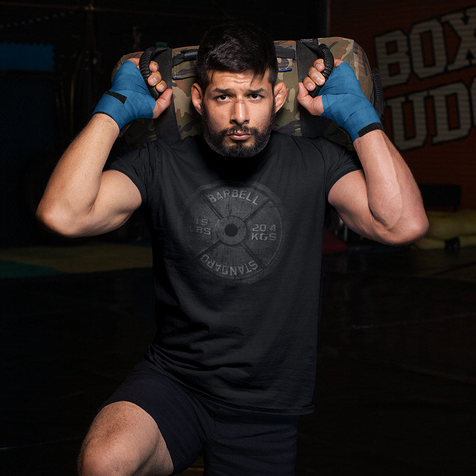 man at gym wearing a workout shirt featuring a faded barbell. The gym shirts for men are side-seamed for an athletic fit. The gyme apparel makes great gifts for gym lovers.